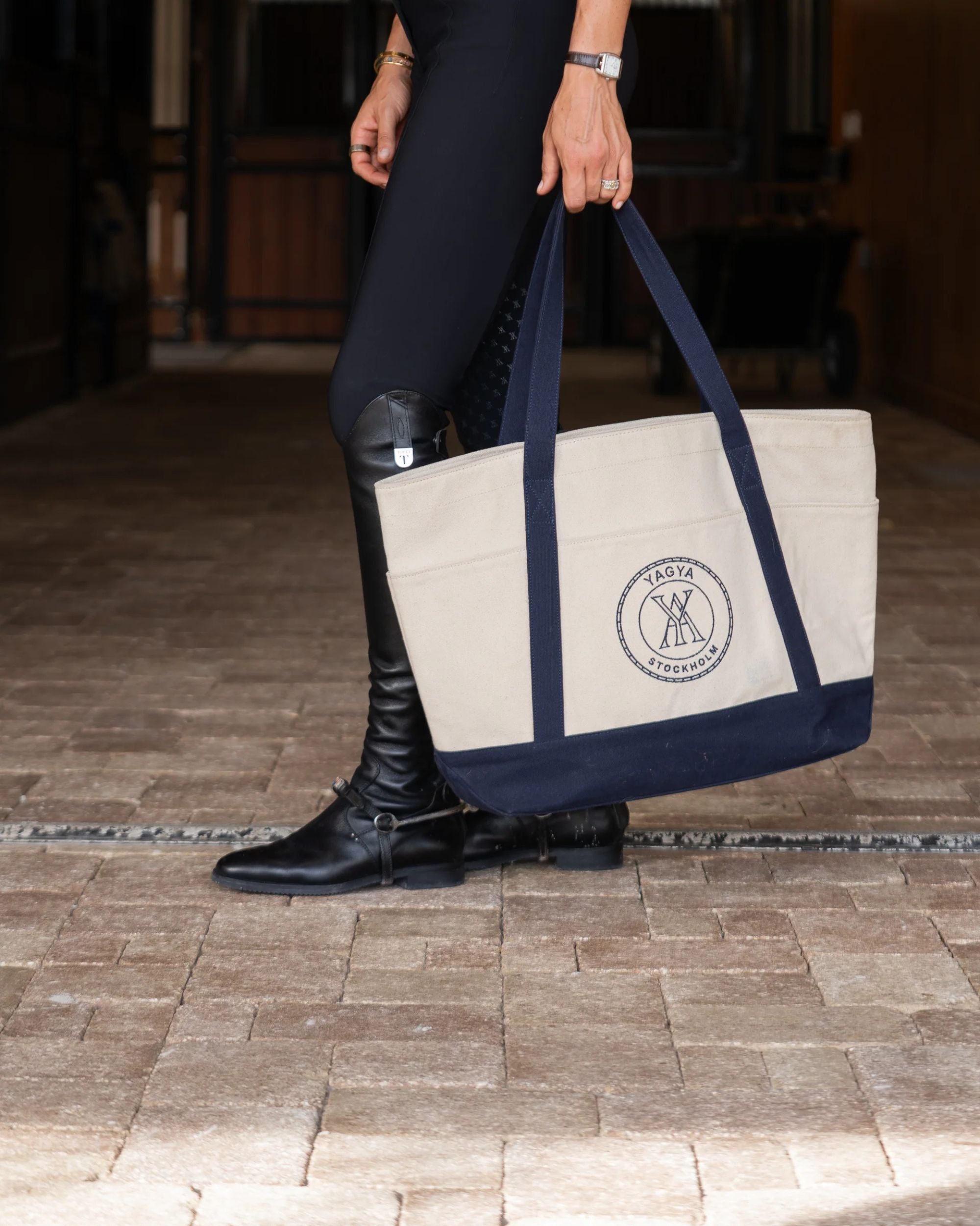 Person holding a beige and navy tote bag with a logo, wearing black riding boots on a brick path.